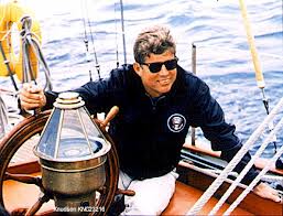 A man in sunglasses sits at the helm of a sailboat, wearing a navy jacket with a circular emblem.