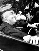 Man smiling while driving a car, wearing a hat and suit.