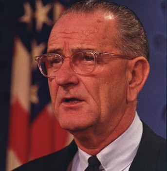 Man with glasses in a suit and tie, speaking with a serious expression. American flag in the background.