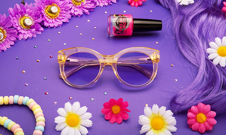 Clear round glasses with a pink nail polish bottle, purple flowers, candy bracelet, and multicolored beads.