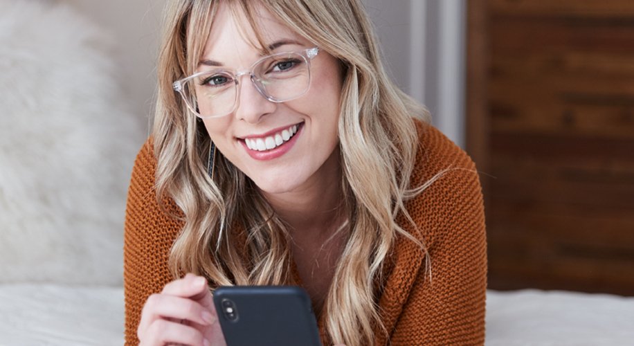 Woman wearing clear-framed glasses and an orange sweater, smiling while looking at a smartphone.