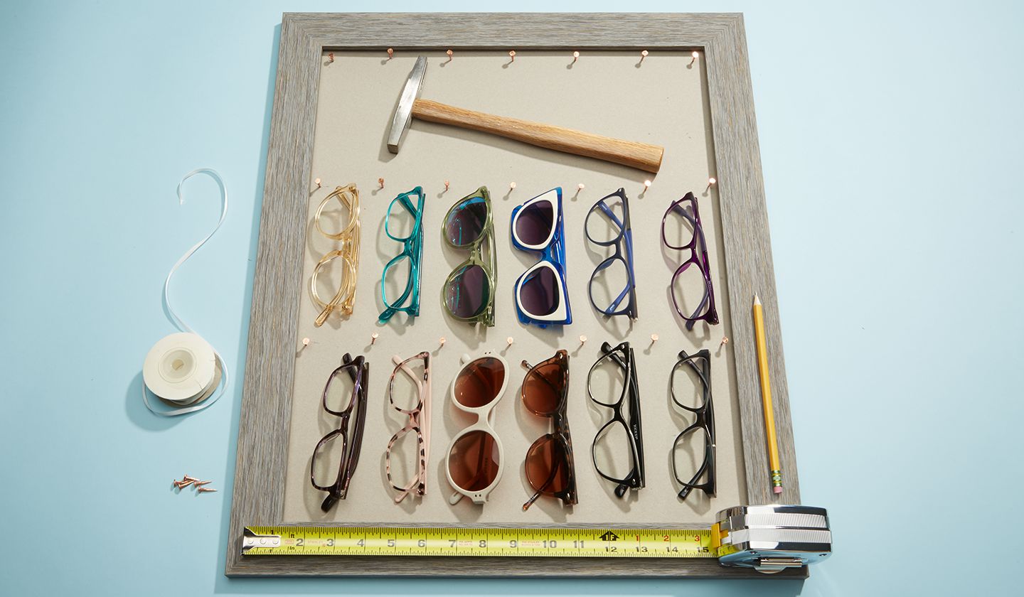 Eyeglasses of various colors and styles arranged in a wooden frame, with a hammer and measuring tape.