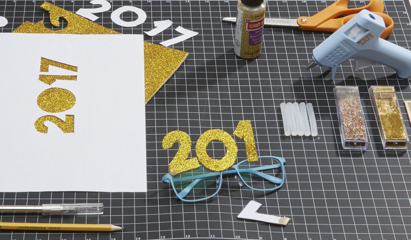 DIY craft project with glittery 2017 cutouts, craft glue, scissors, and blue glasses.