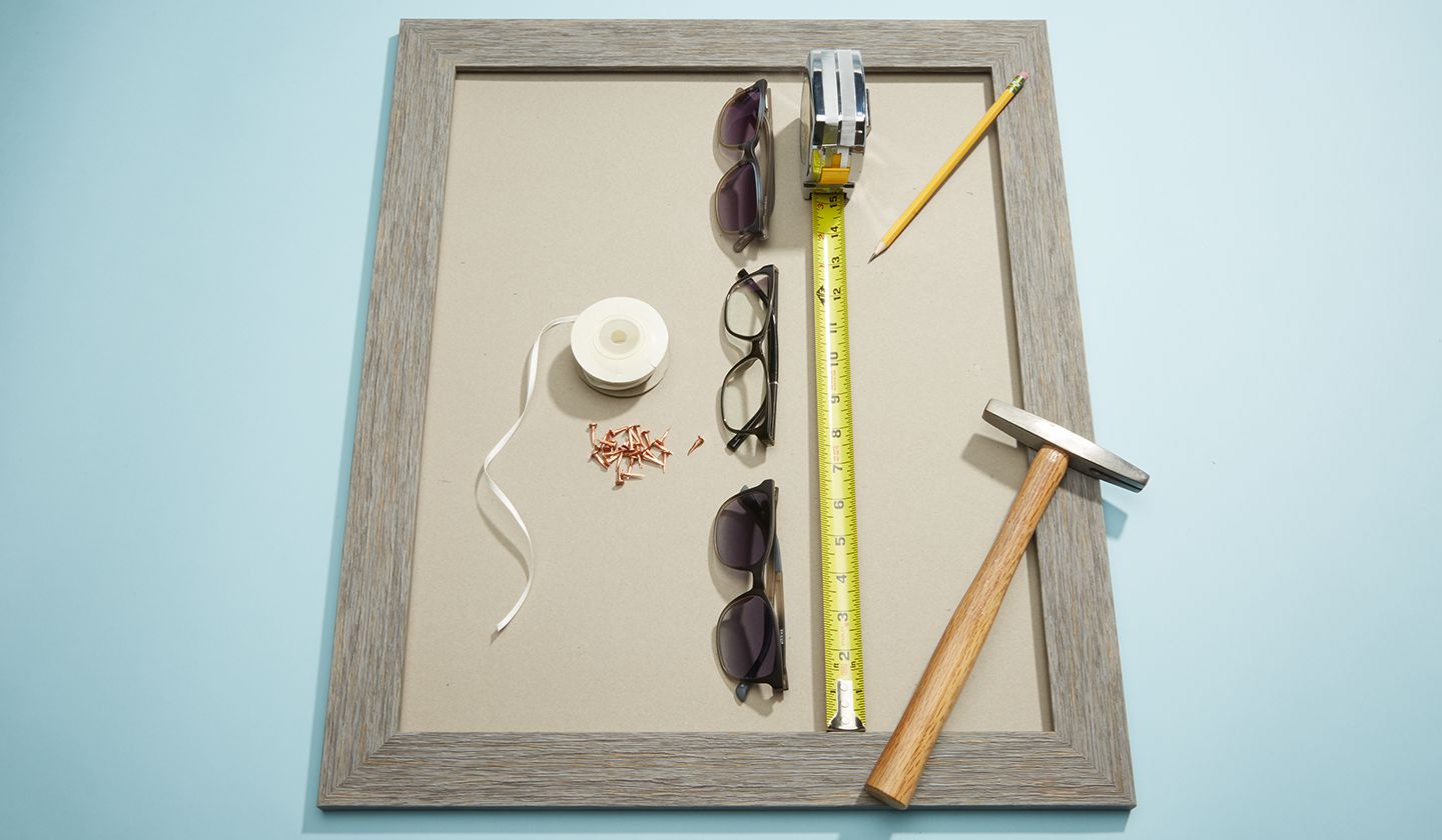 Glasses, tape, measuring tape, pencil, ribbon, tacks, and hammer on a wooden frame on a blue surface.