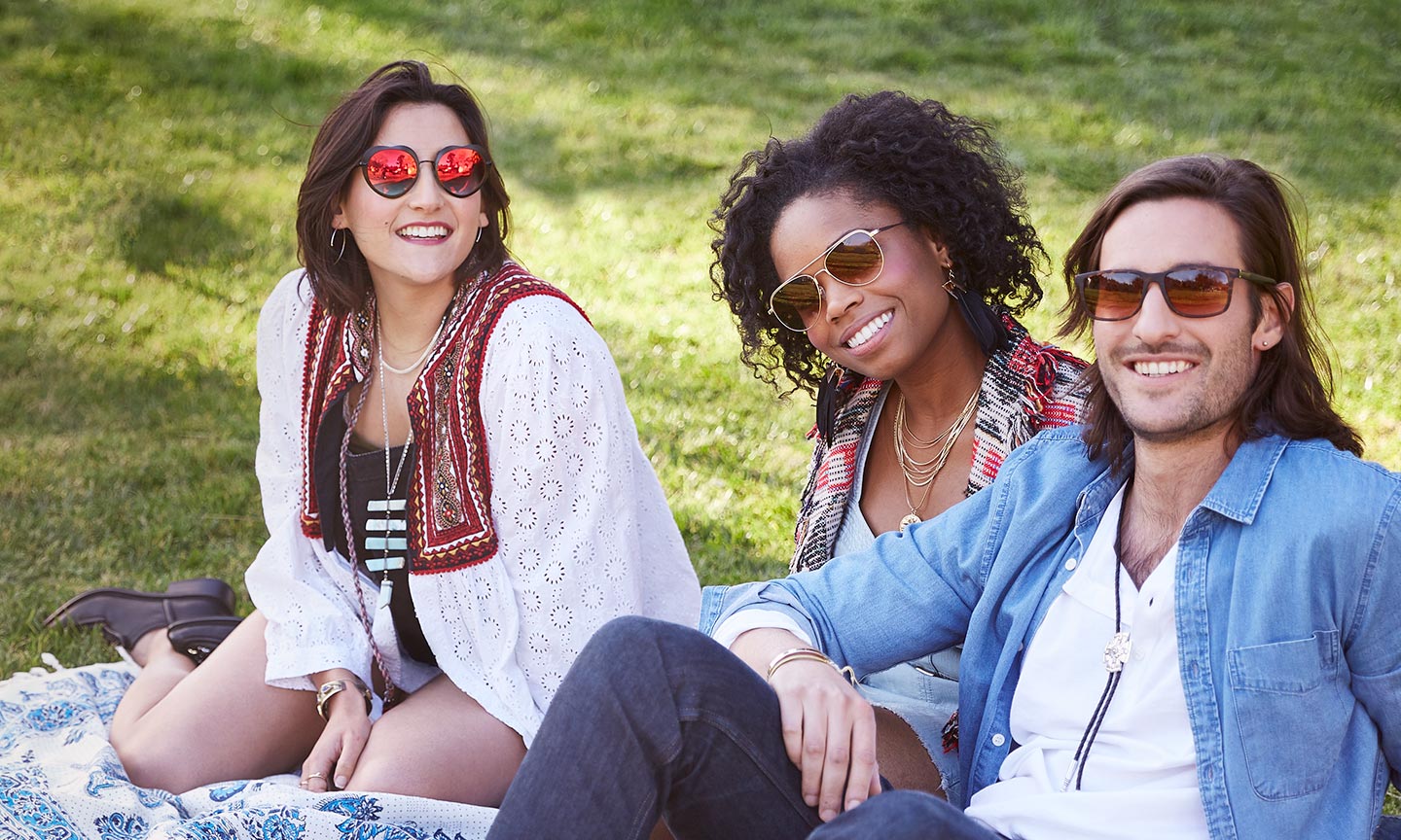Three people wearing stylish sunglasses, sitting on a picnic blanket outdoors and smiling.