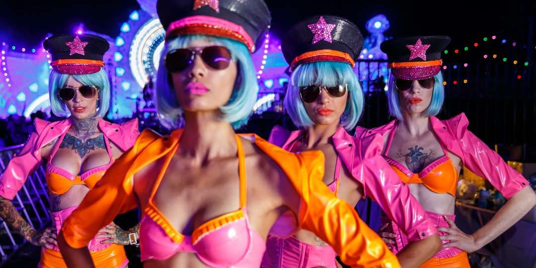 Four women wearing matching bright pink and orange costumes with blue wigs, sunglasses, and star-decorated hats.