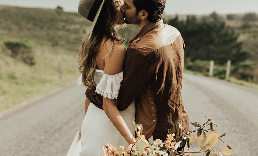 Man in brown shirt kissing a woman in a white dress holding a bouquet of flowers on a rural road.