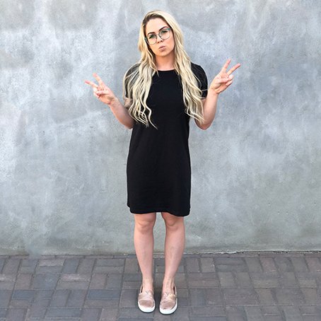 Woman wearing a black knee-length dress, glasses, and slip-on shoes, making peace signs with both hands.