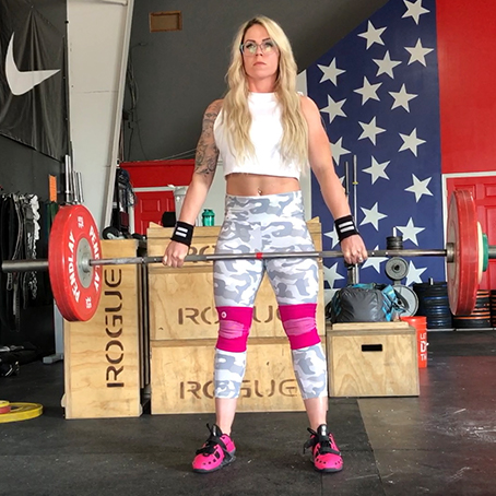 Woman lifting a barbell with red weights while wearing camo leggings, a white crop top, and pink shoes.