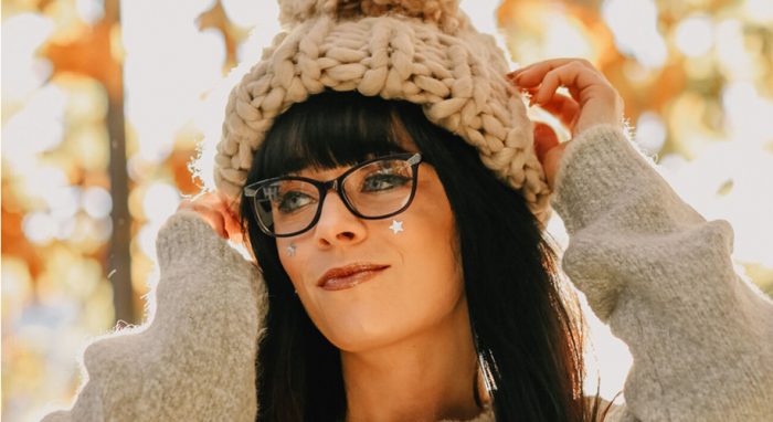 Woman wearing black-rimmed glasses, a chunky knit beanie with pom-pom, and a cozy gray sweater.