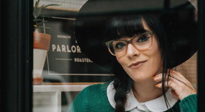 Woman with transparent glasses and wide-brim hat wearing a green sweater, text in the background: PARLOR.