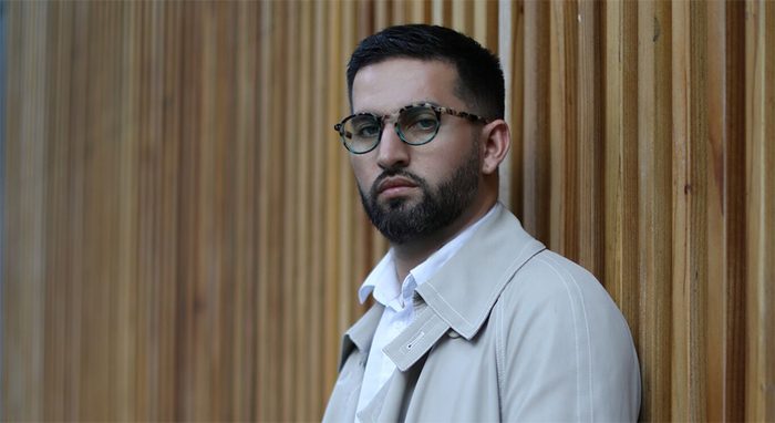 Man wearing tortoiseshell round glasses and a beige trench coat.