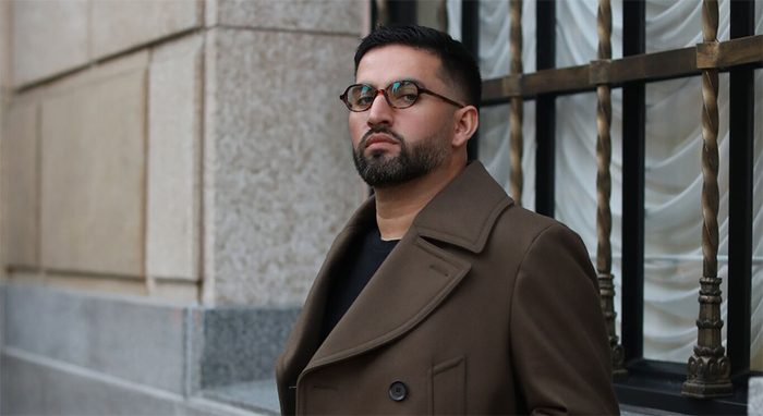 Man wearing black-rimmed glasses and a brown double-breasted coat with notch lapels.