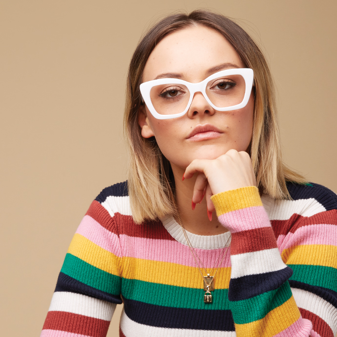 Woman wearing white cat-eye glasses, multi-colored striped sweater, gold necklace with a pendant.