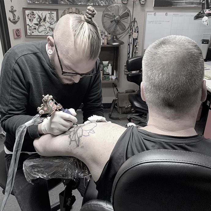 Tattoo artist creating a flower tattoo on a man&#039;s upper arm.