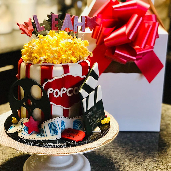 Red and white striped cake designed like a popcorn bucket, with the name &quot;LEAH&quot; on top. Decorative clapperboard reads: &quot;PRODUCED WITH LOVE BY LEAH BAY &amp; FAMILY, TAKES LABS&quot;.