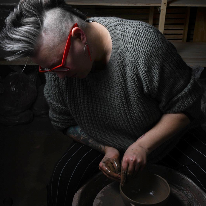 Person crafting a pottery bowl on a pottery wheel, wearing a gray sweater and red glasses.