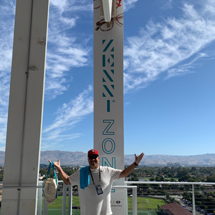 ZENNI ZONE sign on a vertical pillar with a person standing in front, arms raised, blue sky in the background.