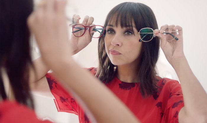 Woman holding red framed glasses and green framed glasses in front of her, deciding which to wear.