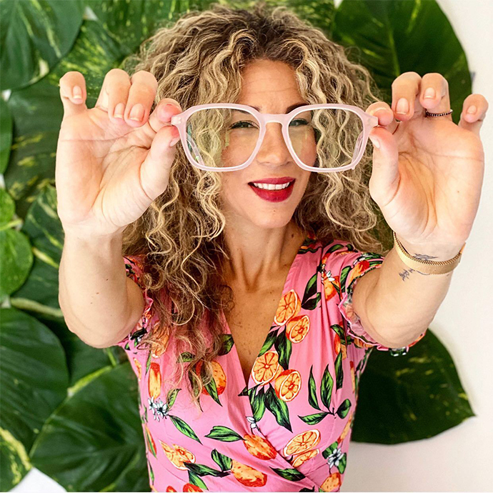 Woman holding pink-rimmed glasses, wearing a colorful floral dress.
