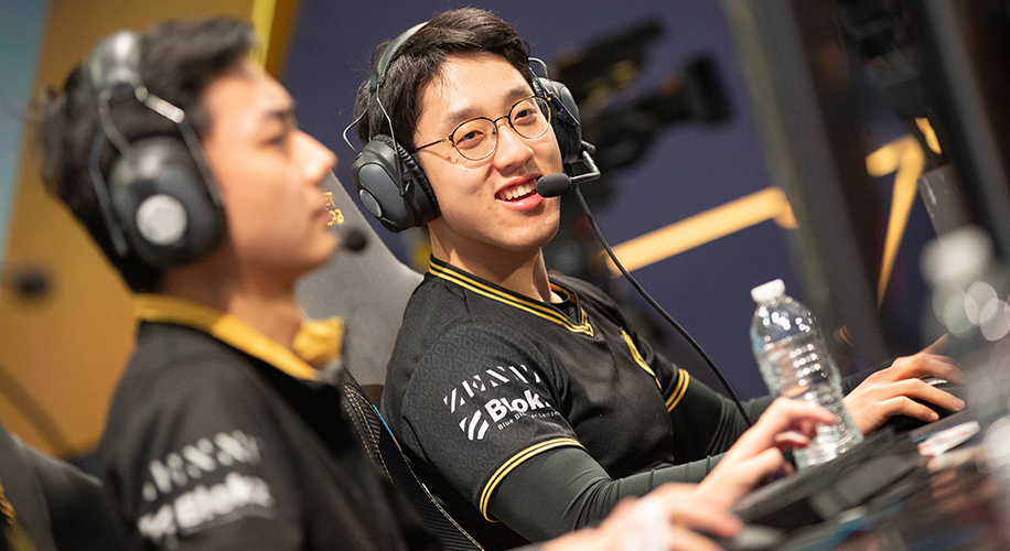 Two gamers wearing black and yellow shirts, headsets, and microphones, smiling while sitting at desktops.