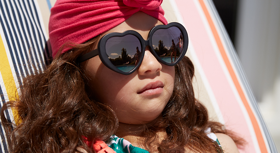 Child wearing black heart-shaped sunglasses and a red headband lying on a striped lounge chair.
