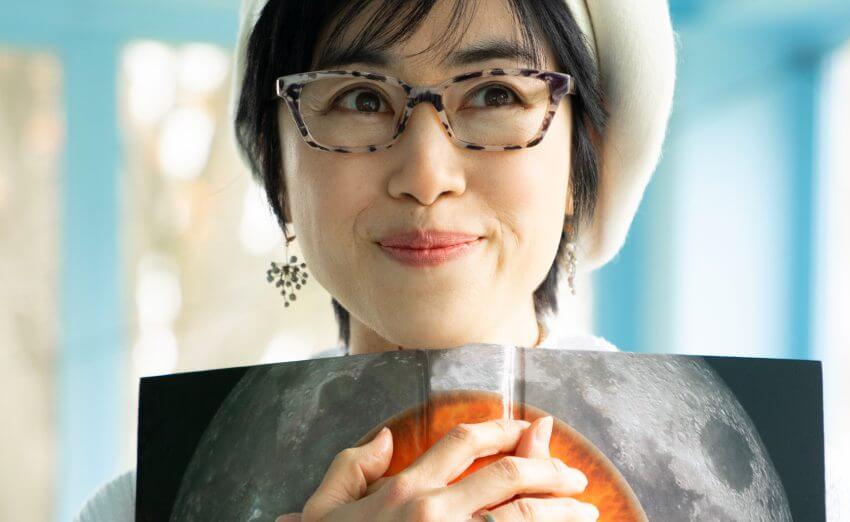 Smiling woman wearing glasses and a beret, holding a book open to an image of the moon and an orange sphere.