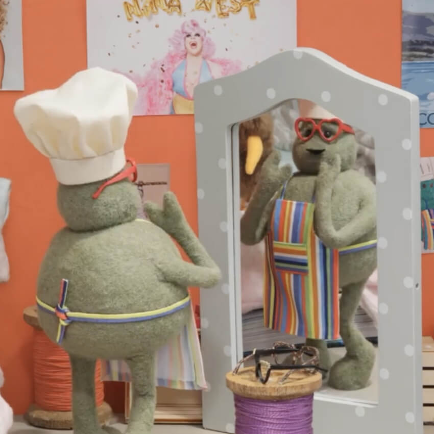 Green plush figure with a chef's hat, red heart-shaped glasses, and a striped apron looking in a mirror.