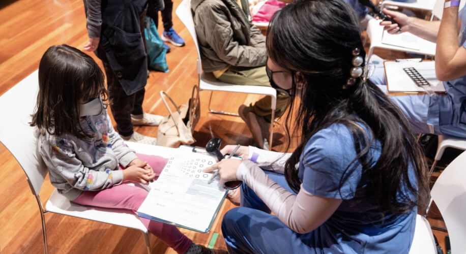 A child wearing a mask, seated and participating in a vision test with a healthcare professional.