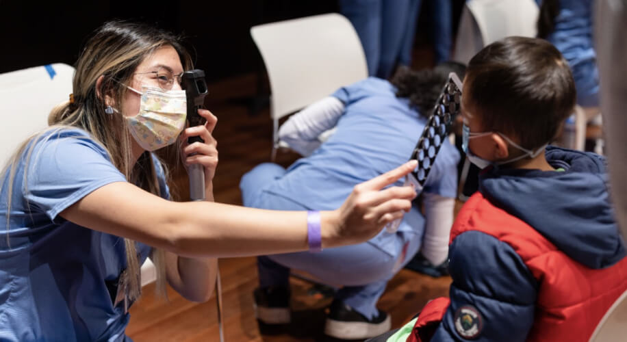 Female healthcare worker performs eye exam on a young boy, holding an occluder in one hand. Both wearing masks.