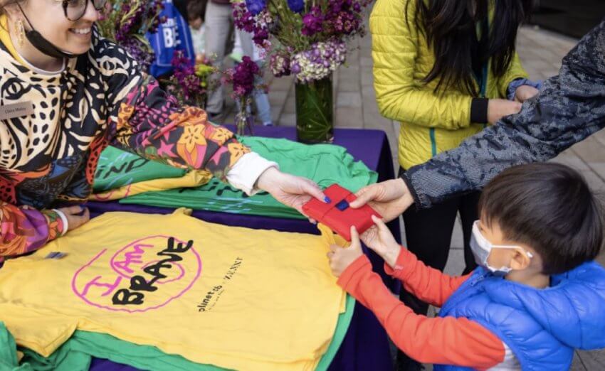 T-shirts with "I AM BRAVE" printed on them and a child holding a red gift bag being handed over.