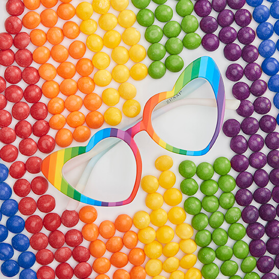 Zenni rainbow eyeglasses with heart-shaped frames. Rainbow candy surrounds the glasses on a white table.