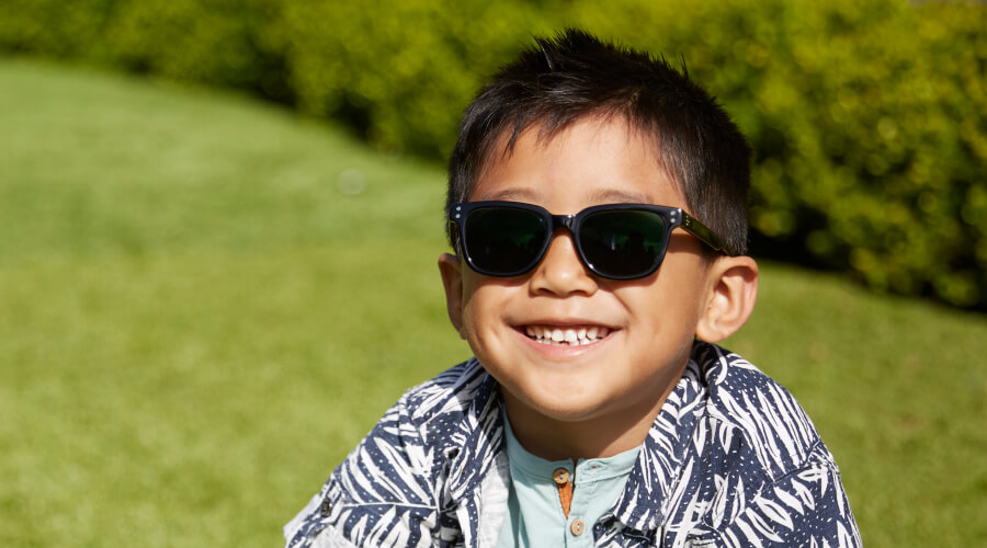 A young boy sits on green field wearing Zenni kids sunglasses. He is wearing a button-up blue and white short sleeve shirt over a light blue t-shirt.