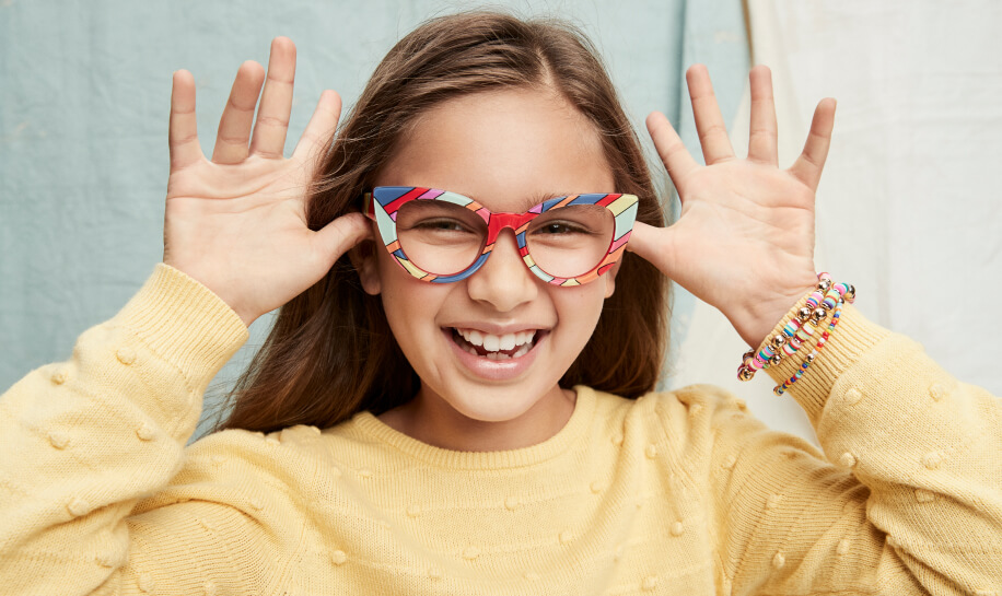 Girl wearing colorful, patterned eyeglasses with a yellow sweater and beaded bracelets, smiling.
