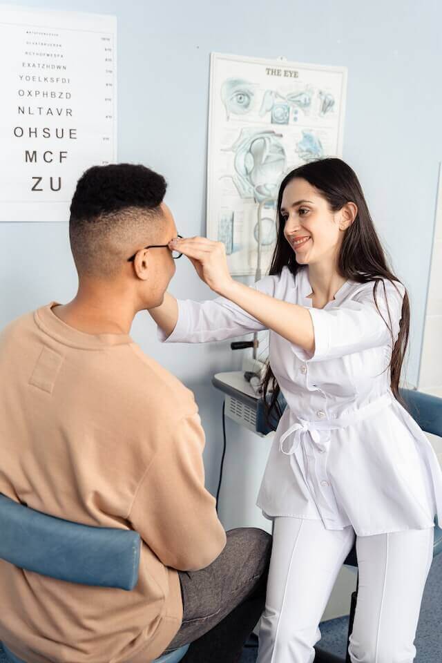 Optometrist examining a patient&#039;s glasses fit, eye chart and eye anatomy poster in the background.