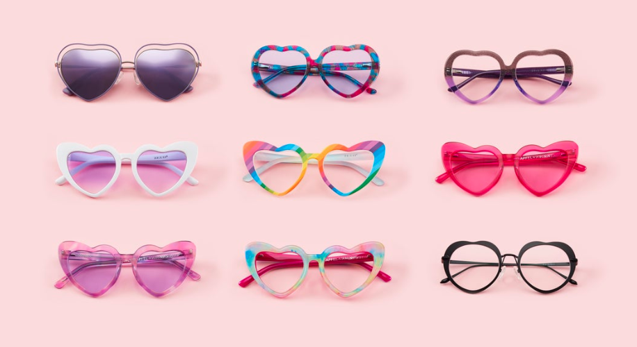 Heart-shaped sunglasses in various colors and designs, arranged in three rows against a light pink background.
