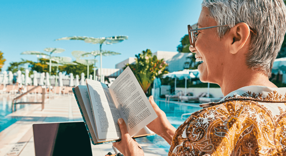 Person reading a book by a pool, smiling, with a clear blue sky and modern shade structures in view.