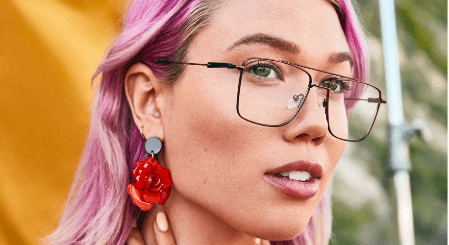 Person with pink hair wearing large square glasses and red rose earrings.