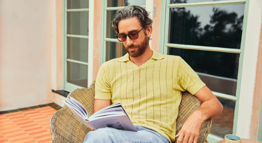 Man in a yellow knit shirt and sunglasses sitting in a wicker chair reading a book.