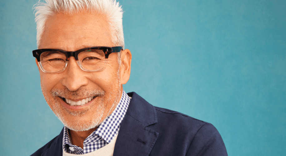 Smiling man wearing black glasses, a blue blazer, and a blue checkered shirt with a white sweater.