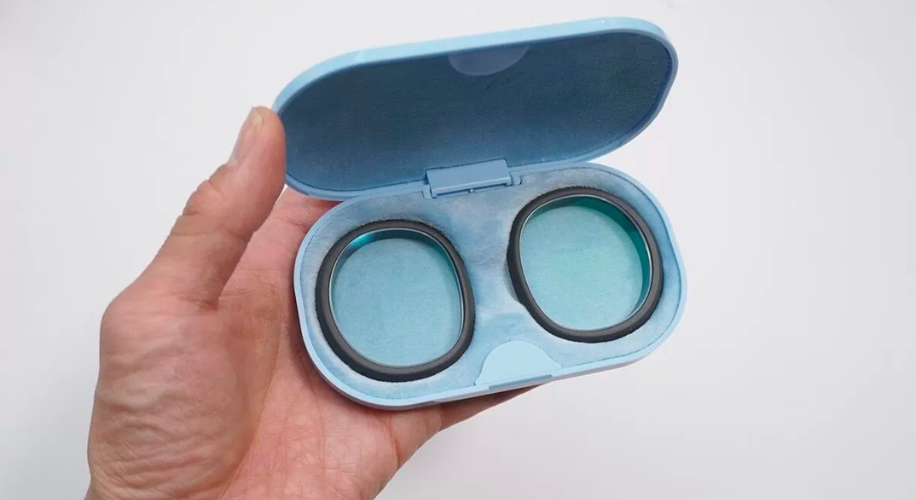 Blue oval case with two greenish-blue eye shadow pans, held by a hand, lid open.