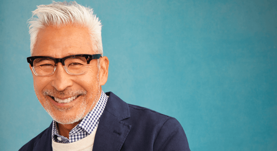 Man with gray hair wearing black glasses, a checkered shirt, and a navy blazer smiles against a blue background.