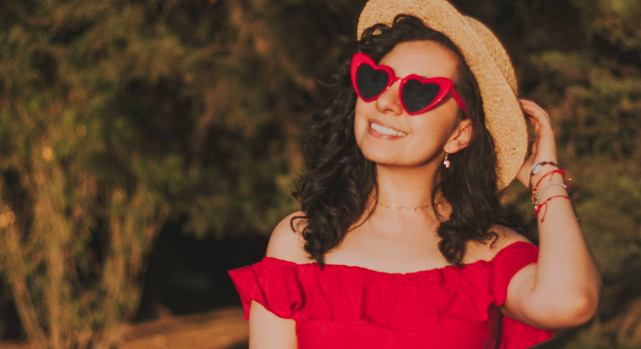 Woman wearing heart-shaped red sunglasses, a straw hat, and an off-the-shoulder red dress.