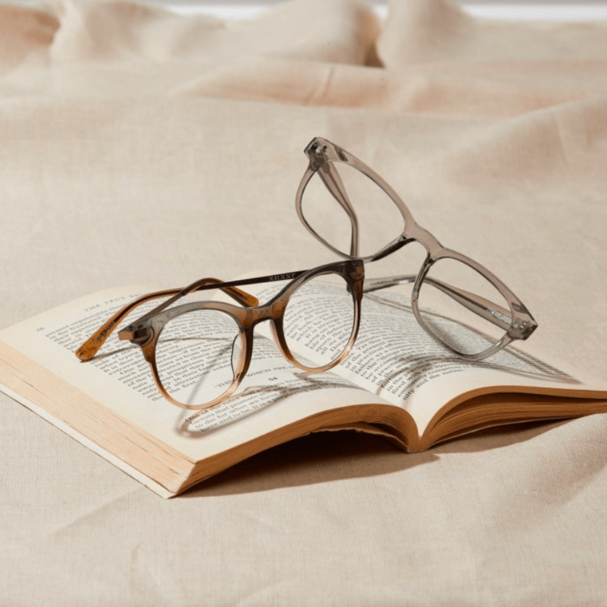 Two pairs of eyeglasses, one with a brown frame and one with a gray frame, lying on an open book.