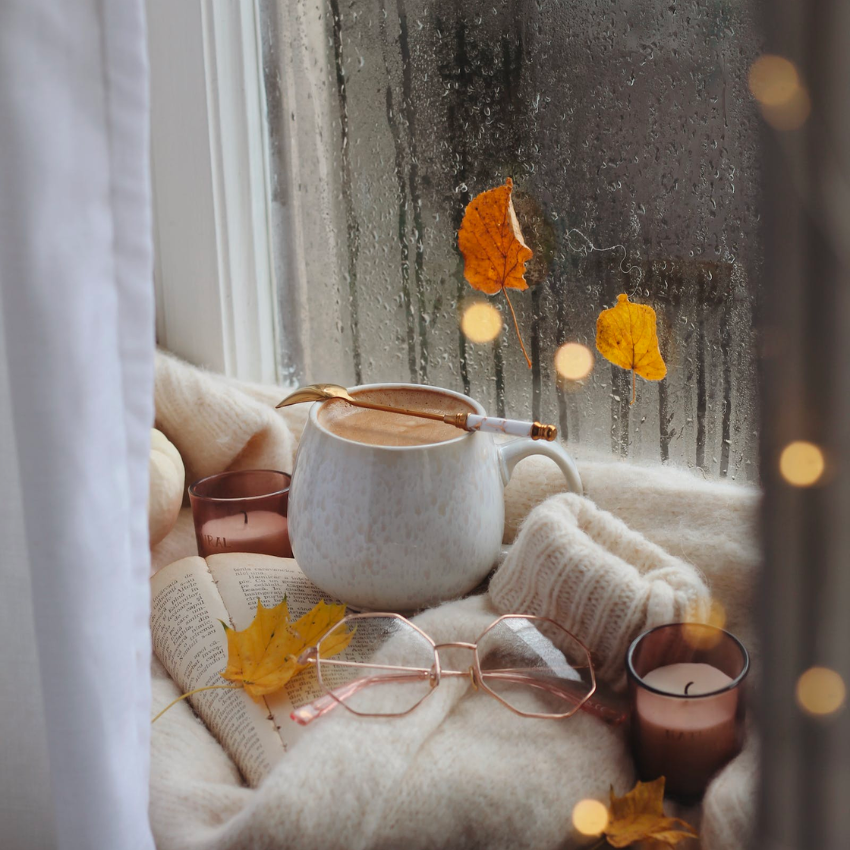 Warm mug of coffee with a gold spoon on a cozy sweater, surrounded by glasses, candles, and a book.