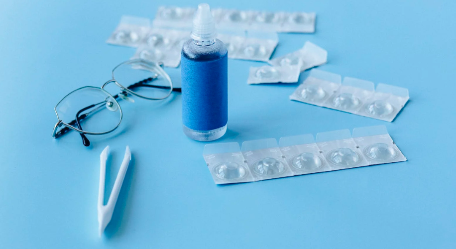 Contact lenses, lens case, tweezers, solution bottle, and eyeglasses on a blue surface.