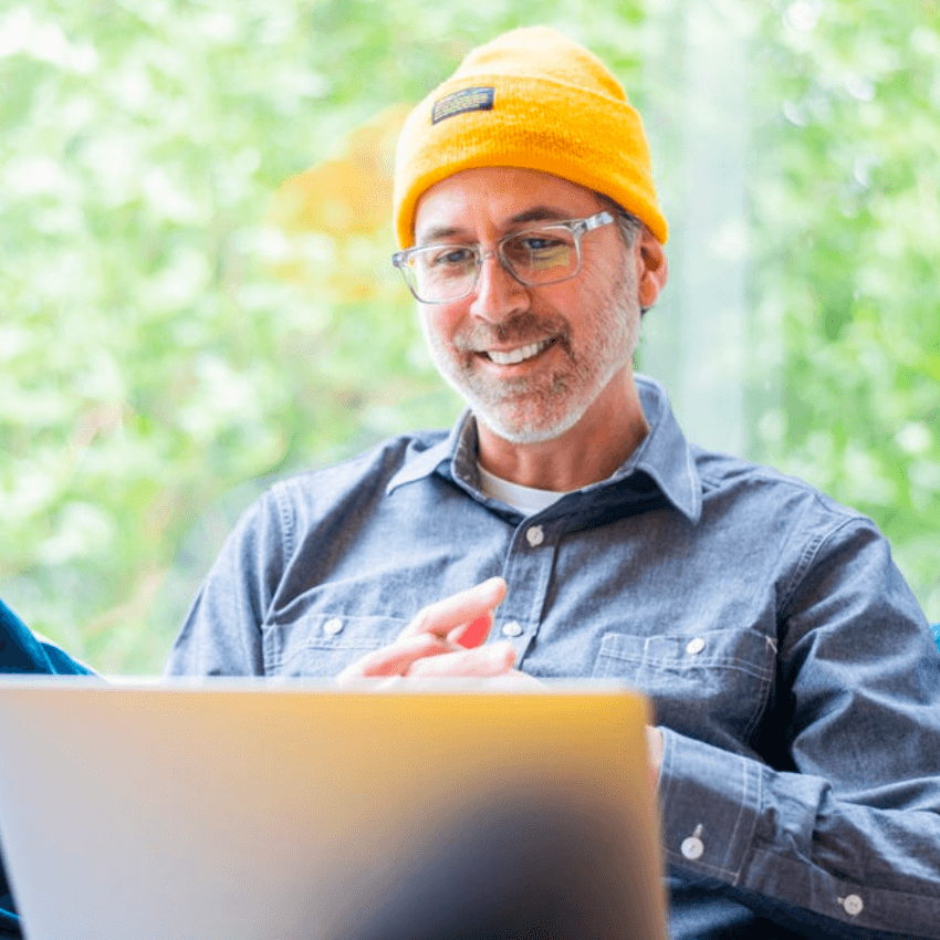 Smiling person wearing a yellow beanie and grey shirt, looking at a laptop.