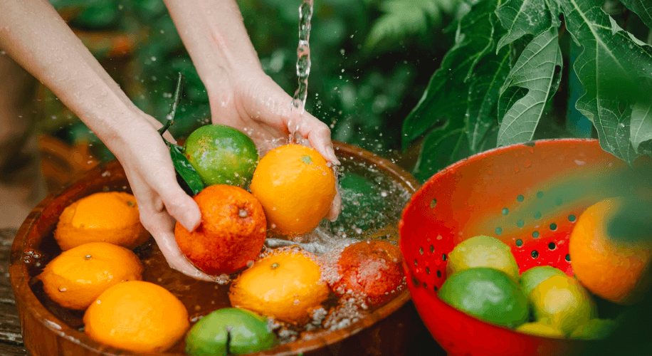 Hands washing assorted citrus fruits in a bowl with running water, next to a basket of limes and oranges.