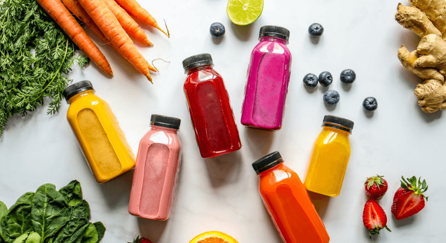 Bottles of colorful fruit and vegetable juices surrounded by fresh produce including carrots and blueberries.
