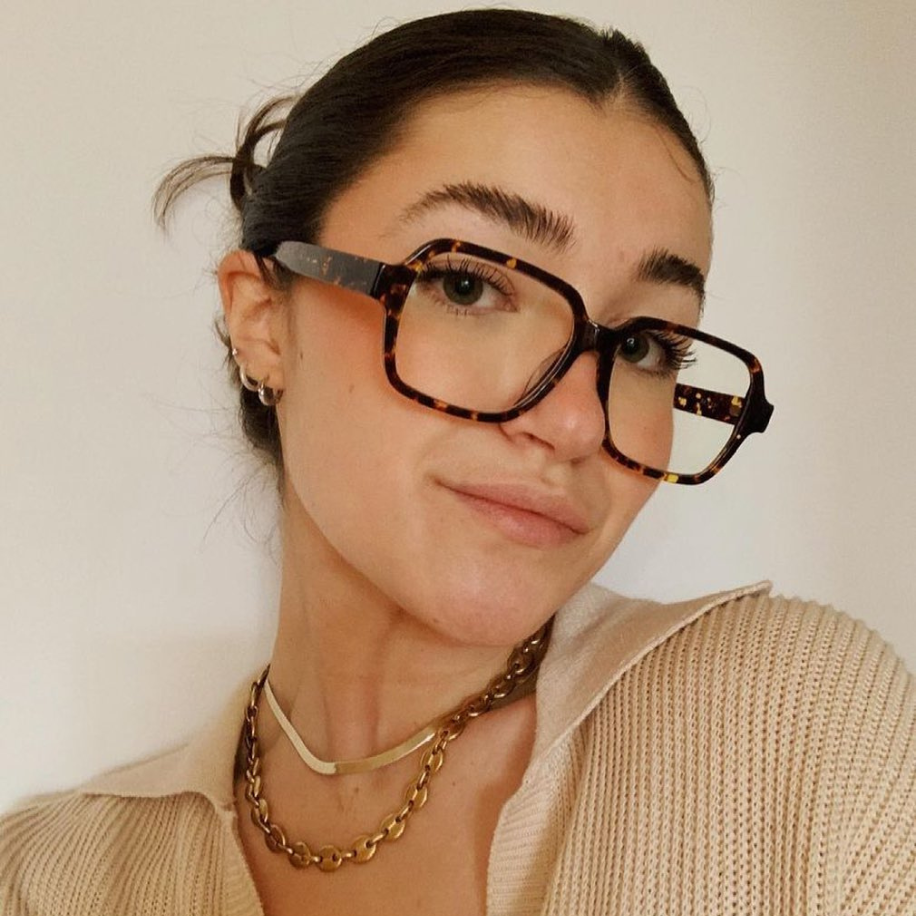 Woman wearing oversized tortoiseshell eyeglasses with geometric lenses and gold jewelry.